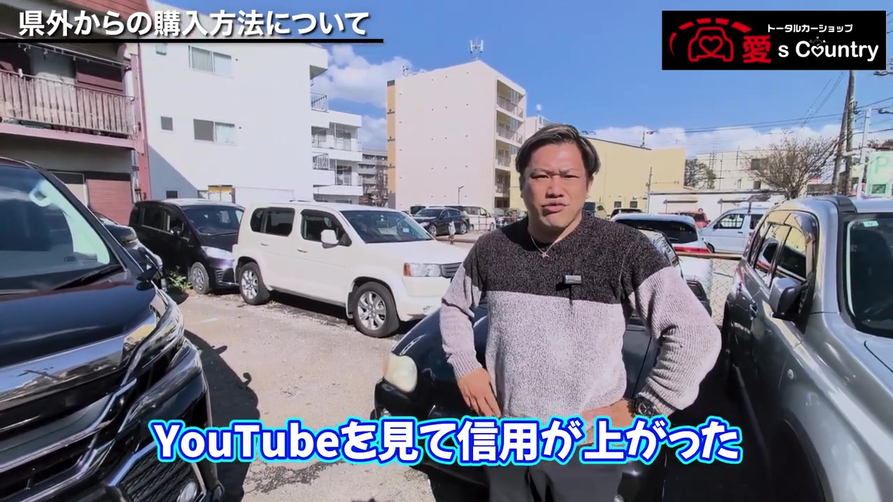 青空の下、車がずらりと並ぶ店頭で県外購入について話す社長